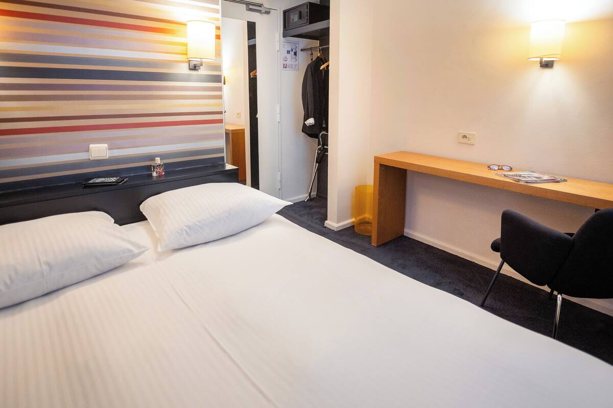 A hotel room with a bed, a desk, and a chair. There is a coat rack with a jacket hanging on it.