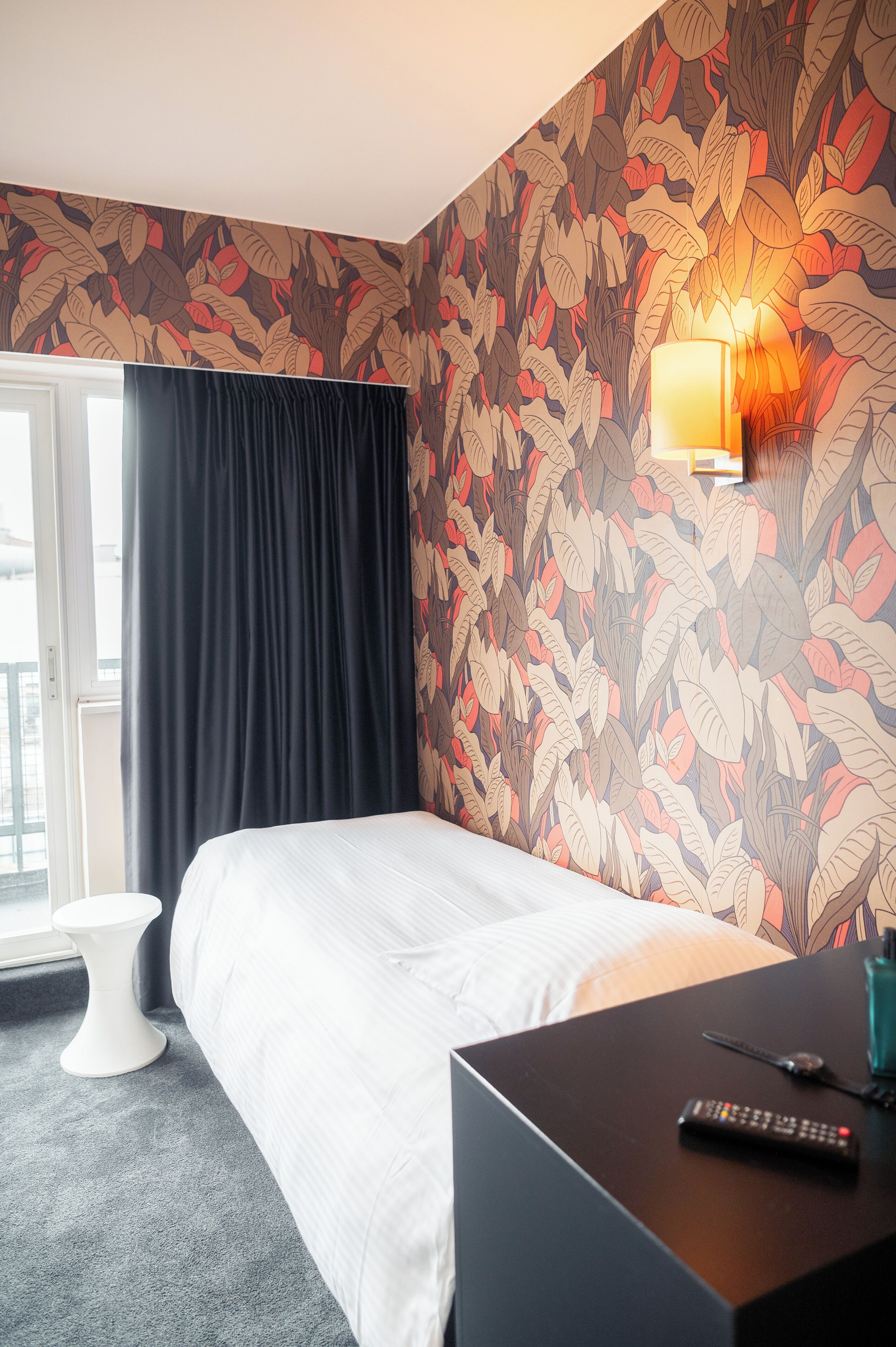 A hotel room with a bed, a bedside table with a remote control, a wall-mounted lamp, and a window with curtains.