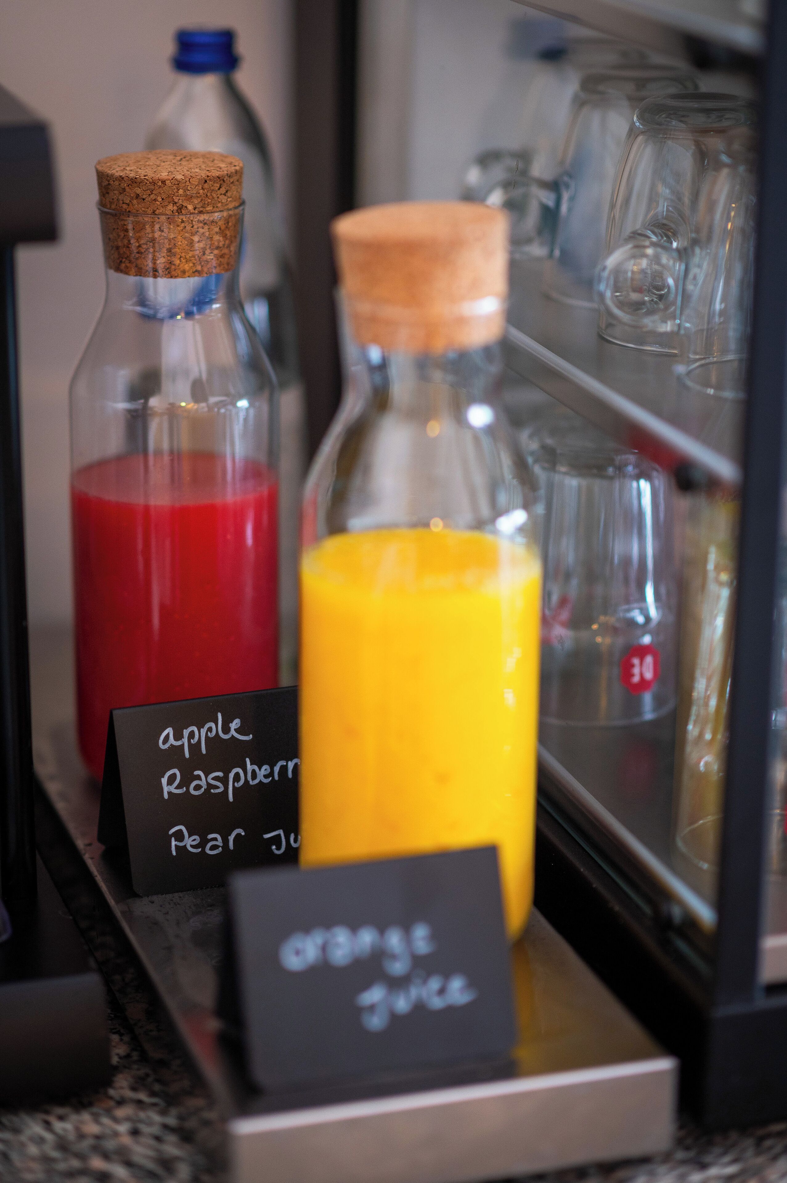 A beverage station with labeled bottles of apple, raspberry, and pear juice, and orange juice.