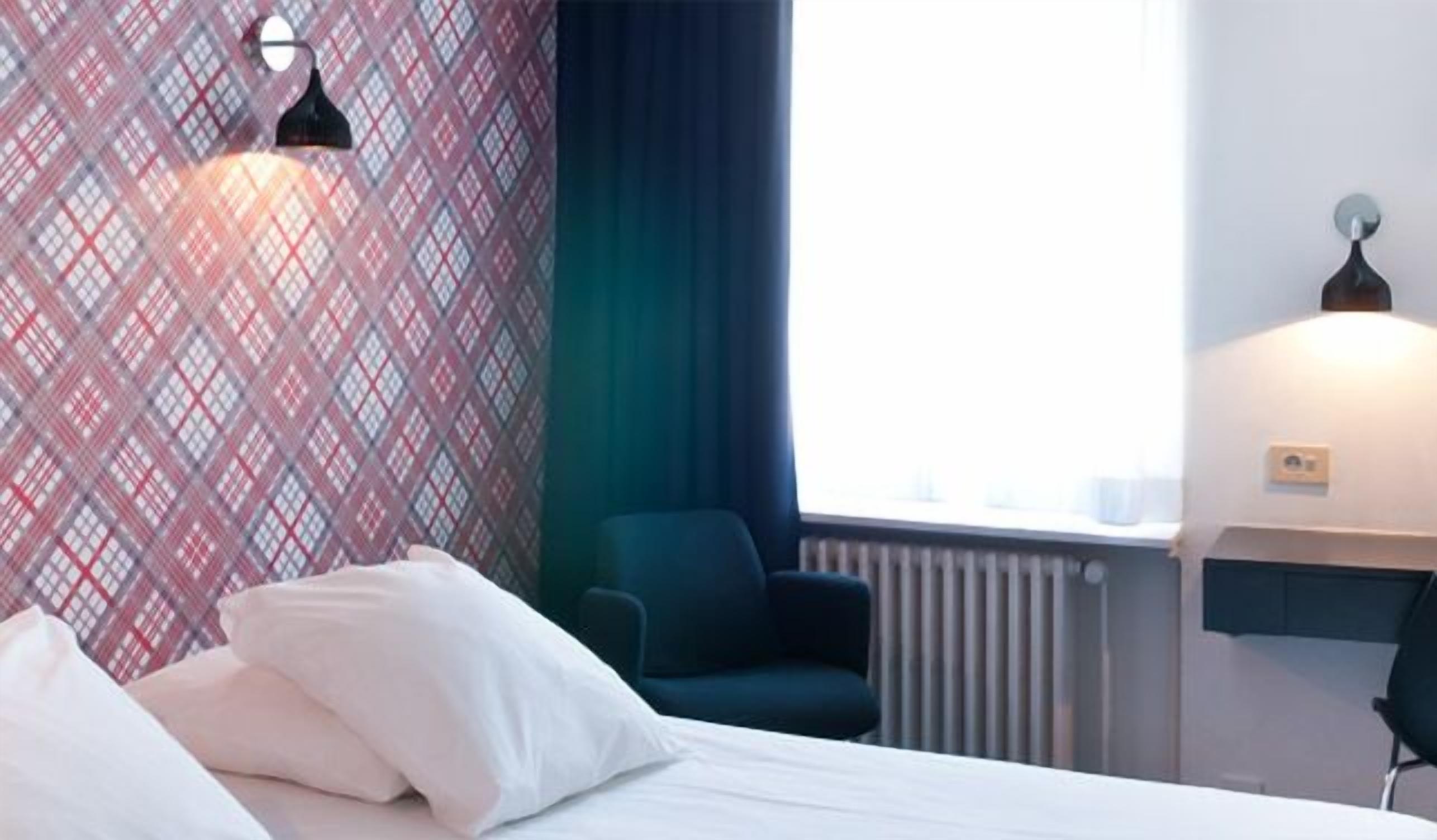 A hotel room with a bed, a chair, a radiator, and a window with curtains.