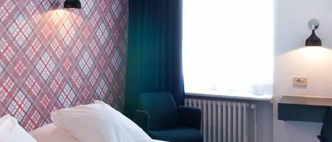 A hotel room with a bed, a chair, a radiator, and a window with curtains.
