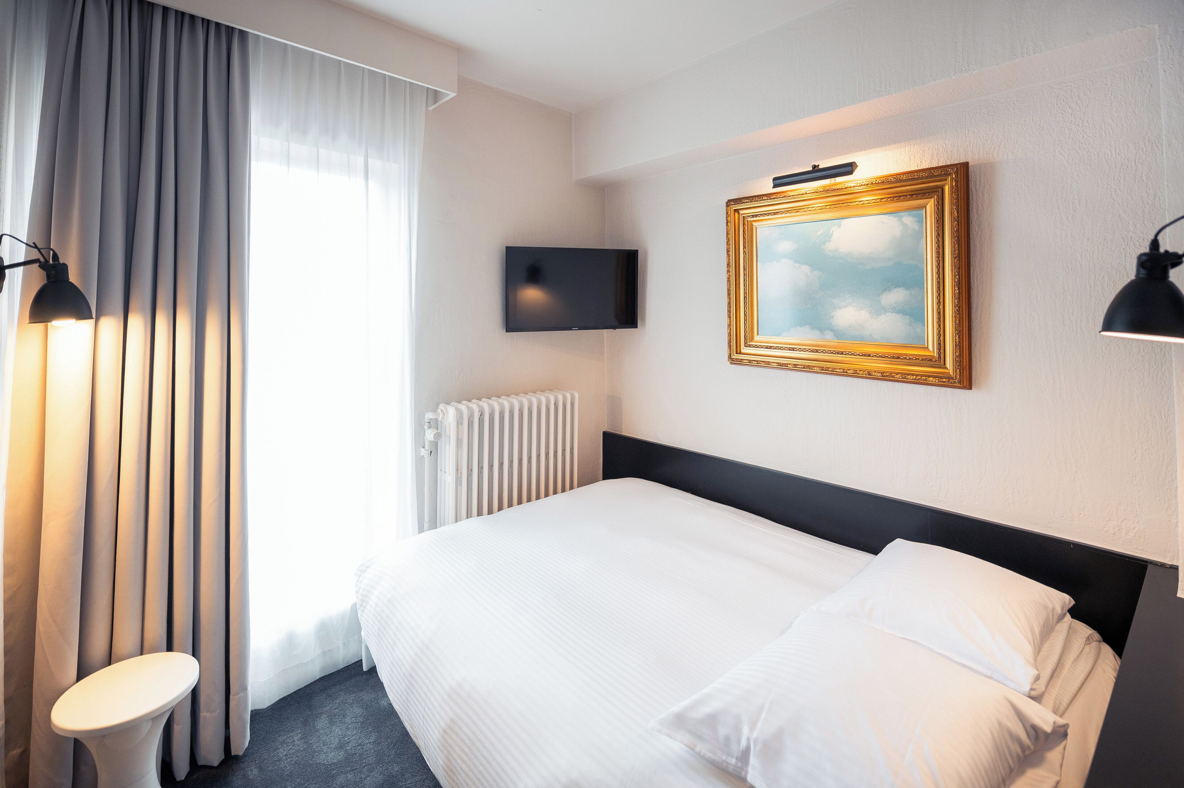 A hotel room with a bed, a television mounted on the wall, a radiator, and a window with curtains.