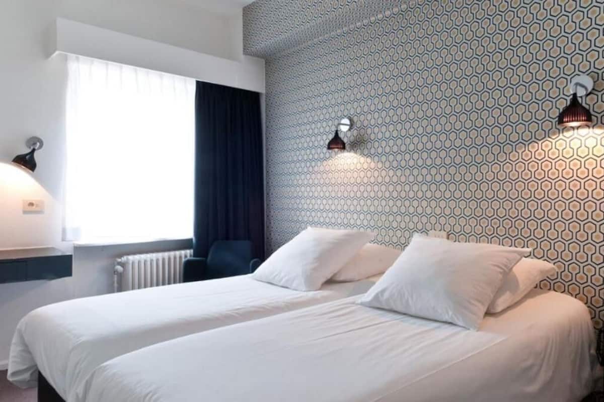 A hotel room with two beds, a window with curtains, a radiator, and patterned wallpaper.