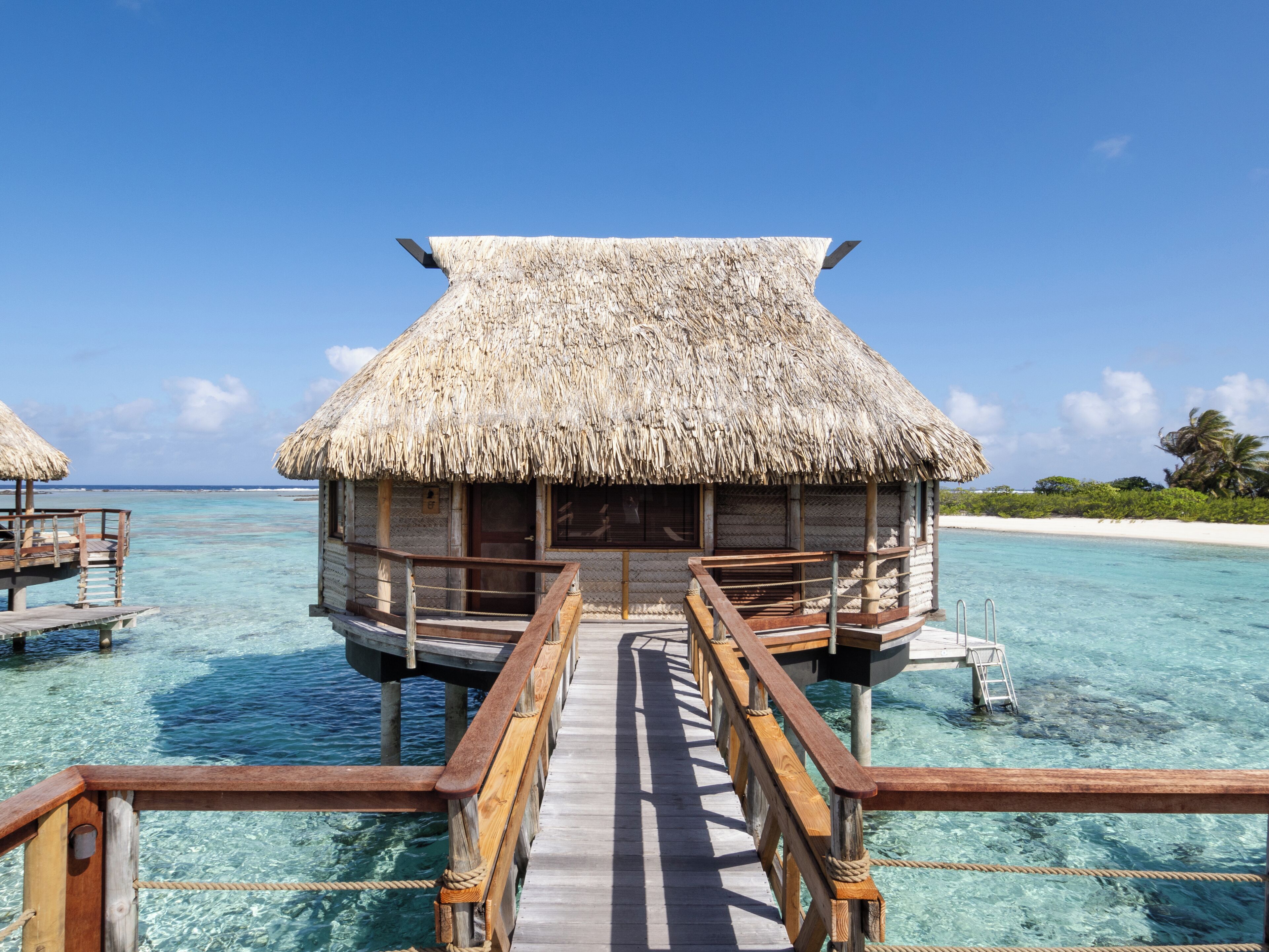 An overwater bungalow with a thatched roof, wooden deck, and clear turquoise water.