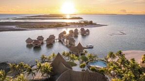Luchtbeeld van bungalows boven water, met een zwembad en palmbomen.