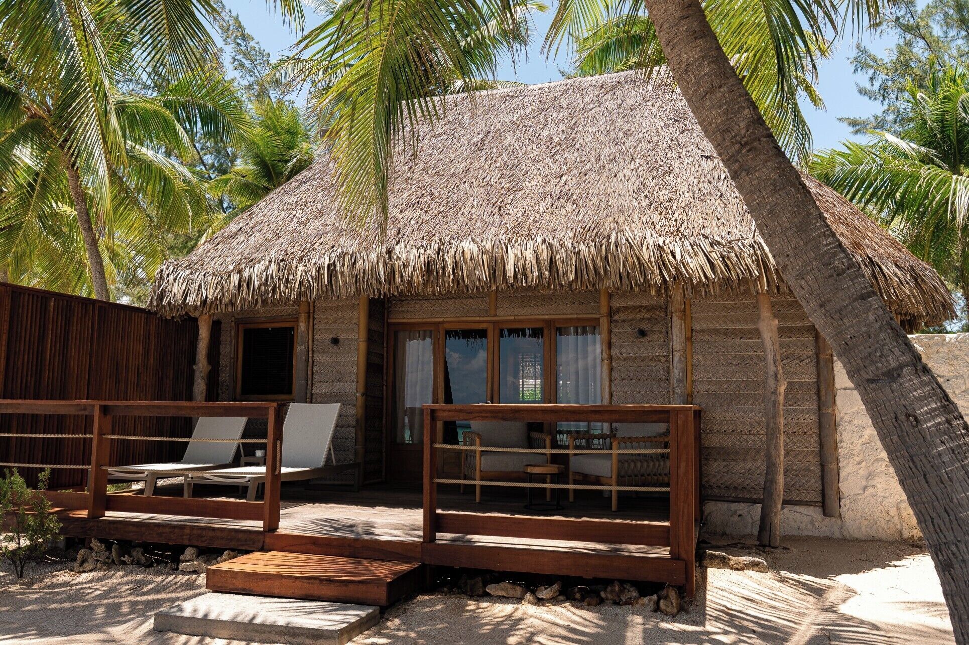 A thatched-roof hut with a porch, wooden furniture, and palm trees.
