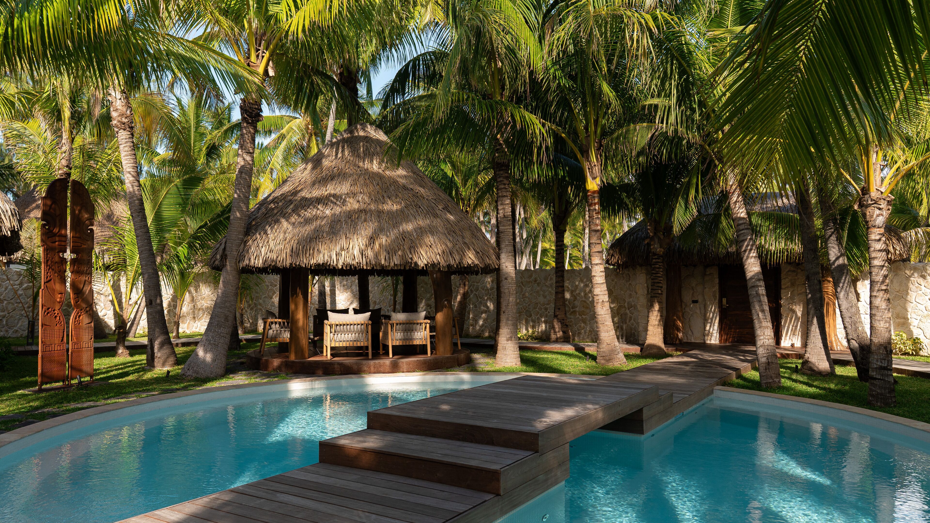 Een zwembadgedeelte met een houten terras, een constructie met rieten dak en palmbomen.
