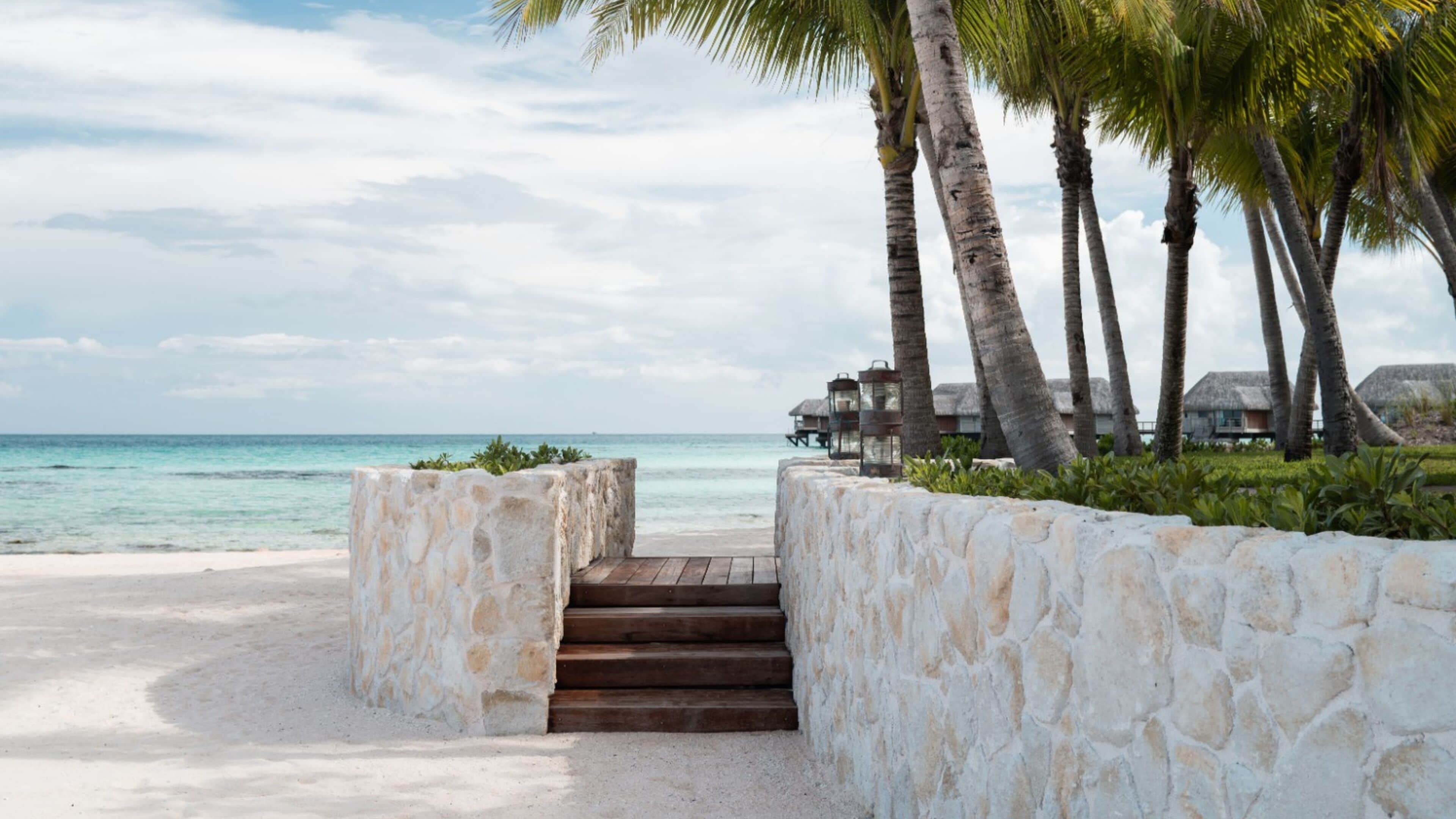 A beach with a stone wall and wooden steps leading to overwater bungalows.