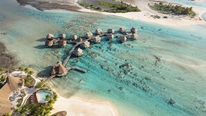 Luchtbeeld van overwater bungalows verbonden door een houten looppad, omringd door helder turquoise water en zandstranden.