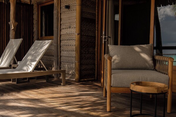 A wooden deck with lounge chairs and a cushioned chair, bamboo walls, and a large window.