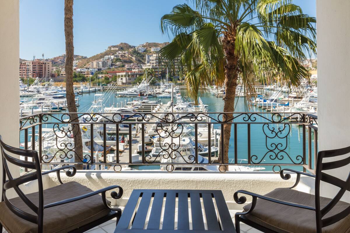 A balcony with two chairs and a table overlooking a marina with boats and a cityscape in the background.