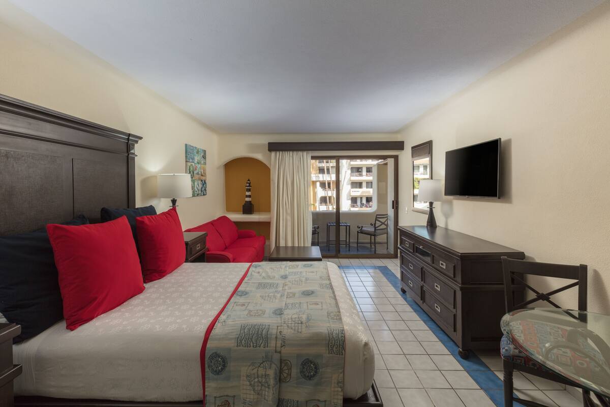 A hotel room with a bed, a desk, a chair, a TV, and a view of the outside balcony.