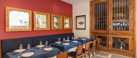 Un coin repas avec une longue table dressée pour le repas, des meubles en bois contenant des bouteilles de vin et des œuvres d’art encadrées accrochées au mur.