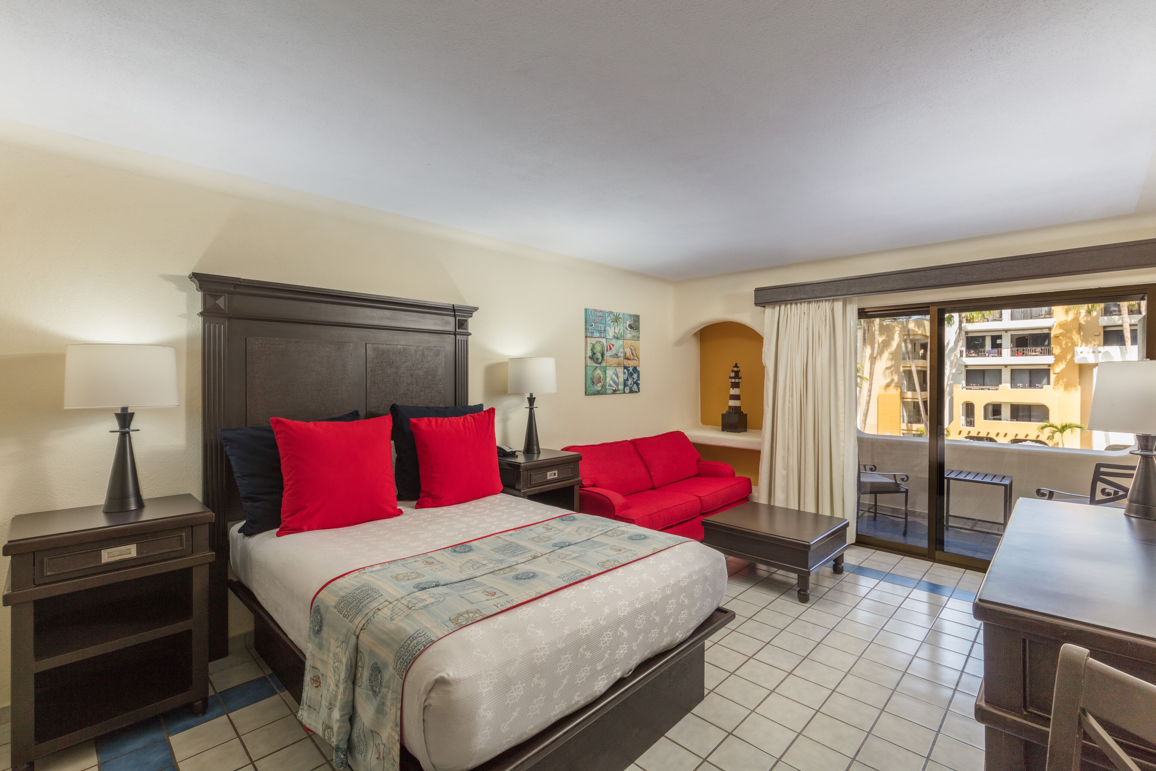 A hotel room with a bed, a red sofa, a dining table, and a balcony with a view of buildings.