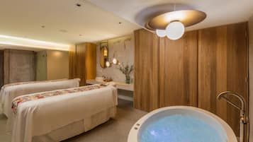 Uma sala de spa com duas macas de massagem, uma banheira grande, painéis de madeira, um espelho e um lustre.