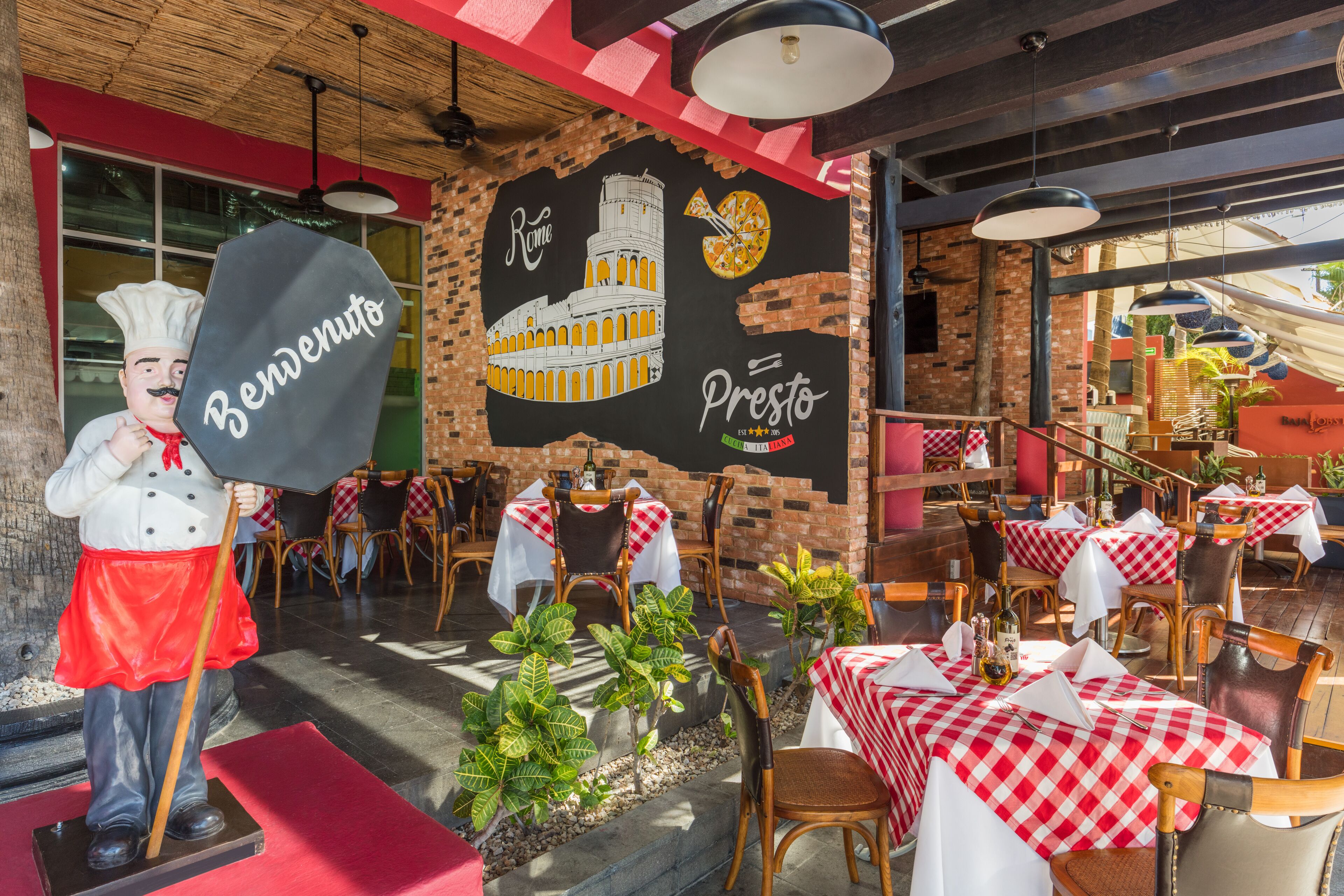An outdoor restaurant with brick walls, red and white checkered tables, and a statue of a chef.