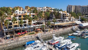 A marina with multiple boats docked, a promenade with shops and restaurants, and a backdrop of multi-story buildings with balconies. - Marina Fiesta Resort & Spa (Cabo San Lucas)