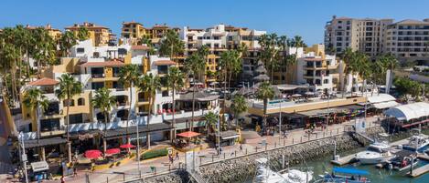 Ein Yachthafen mit mehreren vertäuten Booten, eine Uferpromenade mit Geschäften und Restaurants, sowie im Hintergrund mehrstöckige Gebäude mit Balkonen.