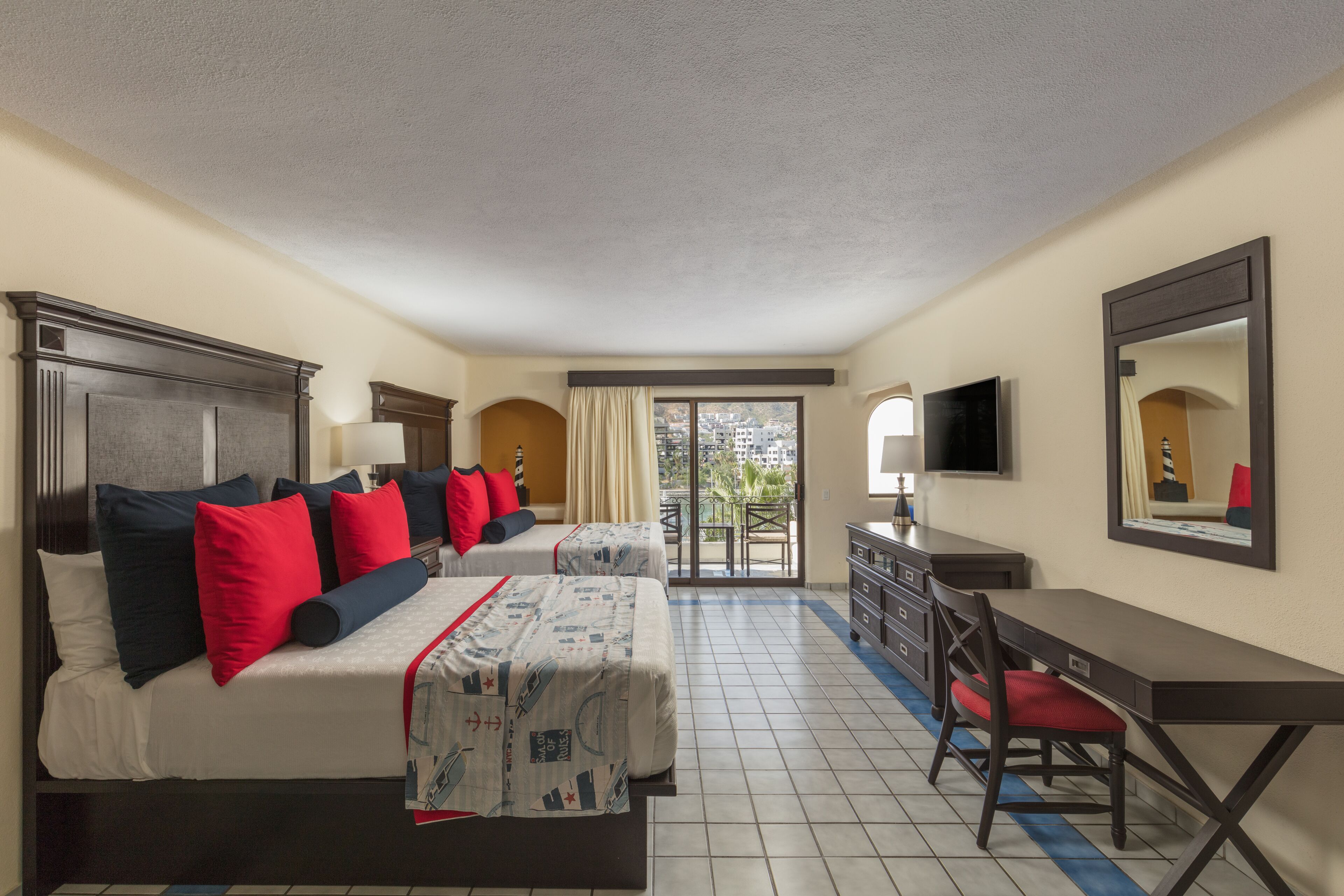A hotel room with two beds, a desk, a chair, a television, and a balcony with a view of buildings.