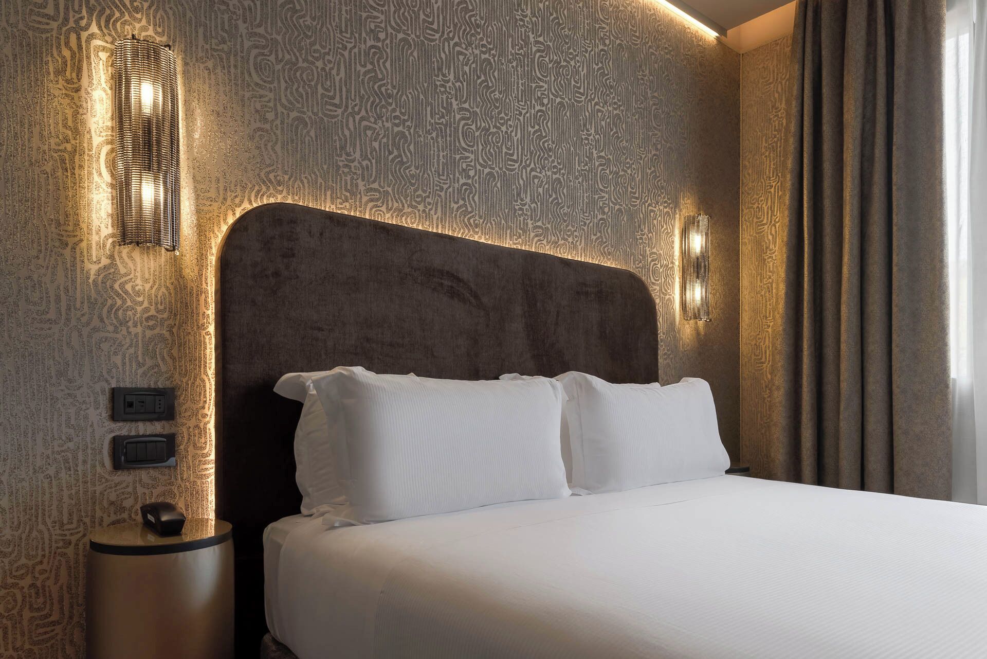 A hotel room with a large bed, a headboard with integrated lighting, a bedside table with a lamp, and a patterned wall.