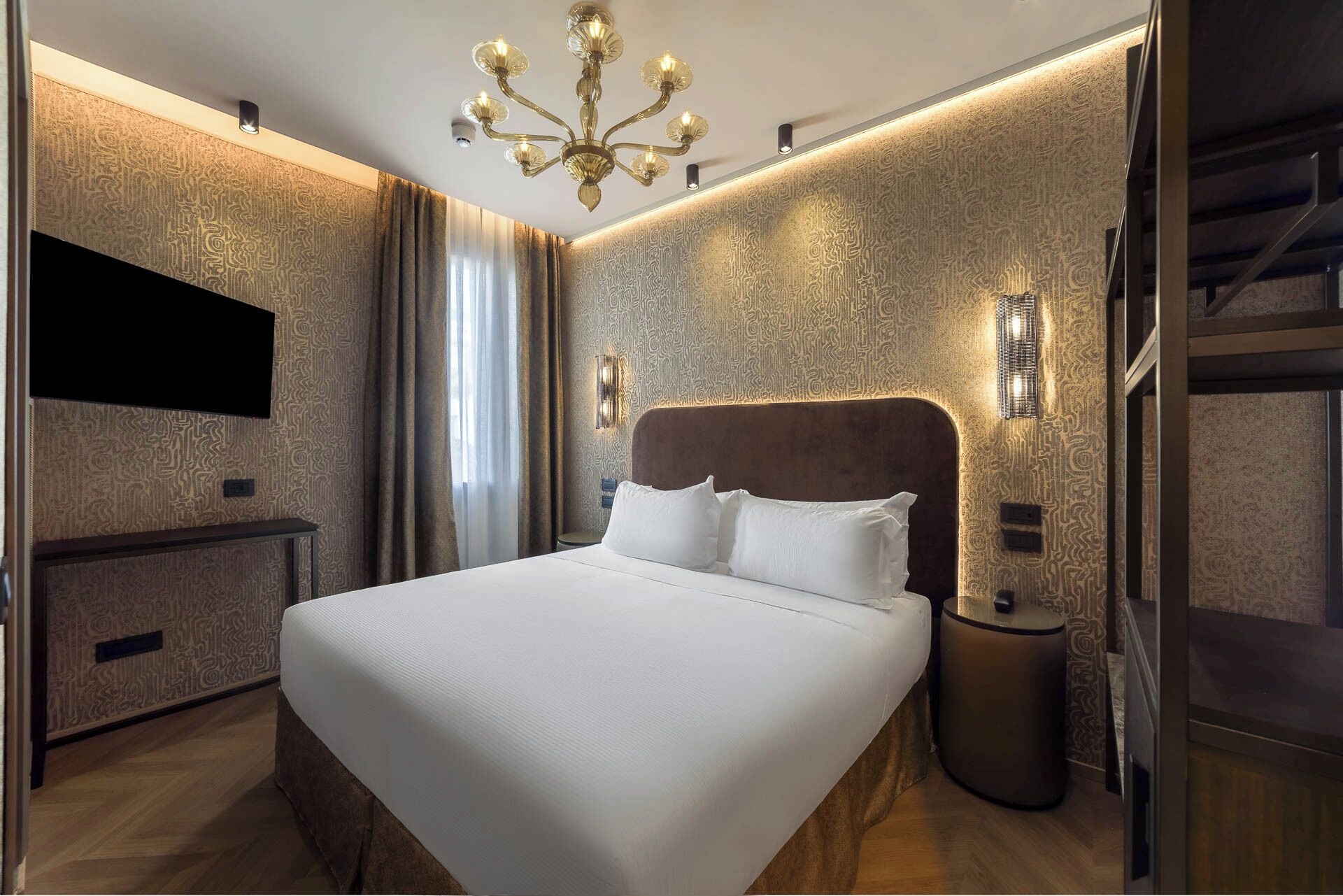 A modern hotel room with a large bed, a TV mounted on the wall, a chandelier, and a staircase.