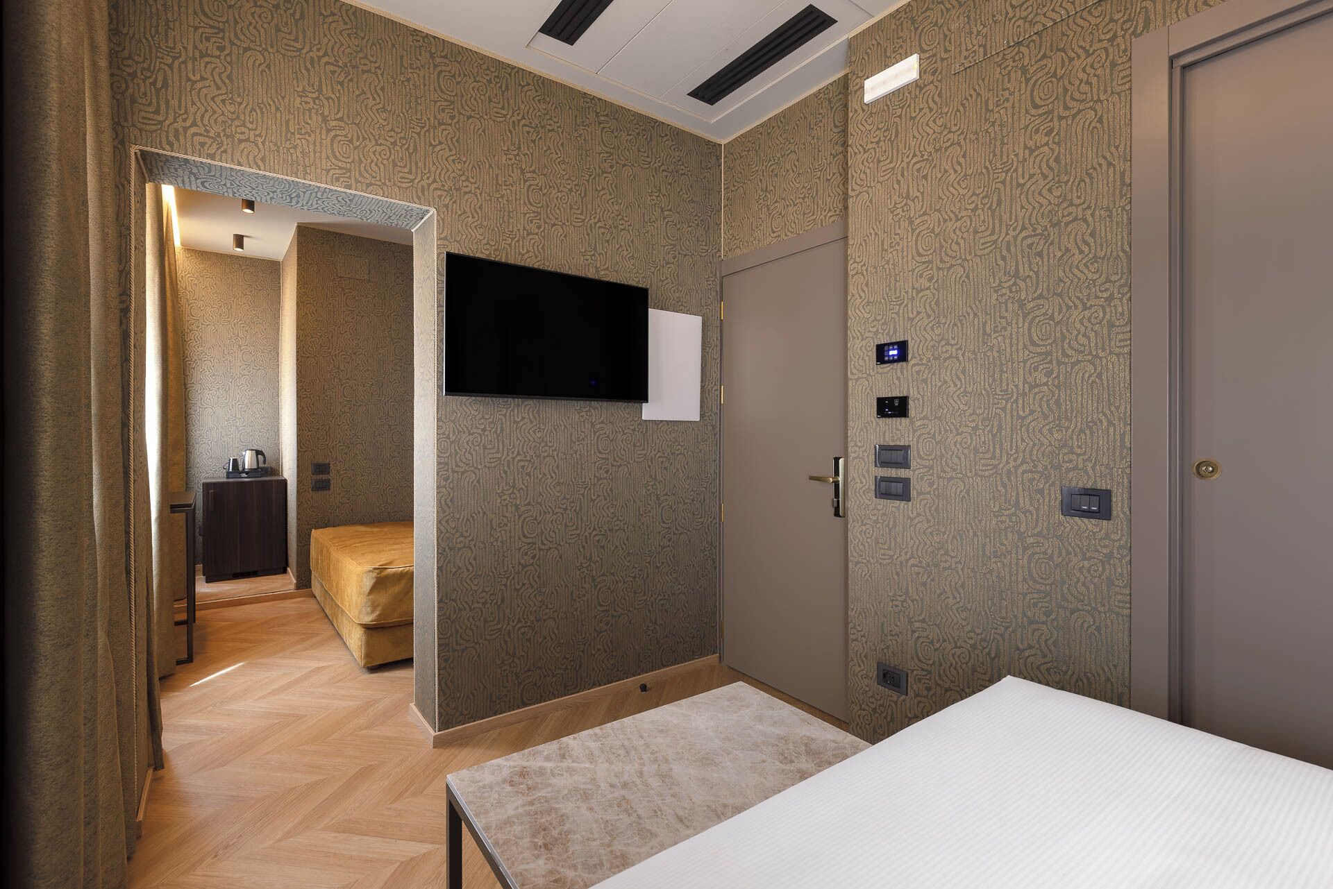 A hotel room with a bed, a television mounted on the wall, a door with a keypad, and a small seating area with a sofa and a table.
