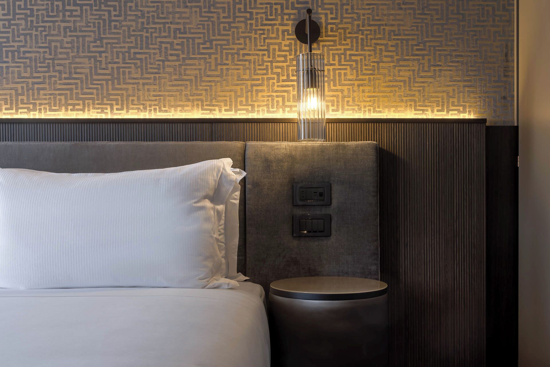 A hotel room with a bed, a bedside table, a wall-mounted light fixture, and a textured wall panel.