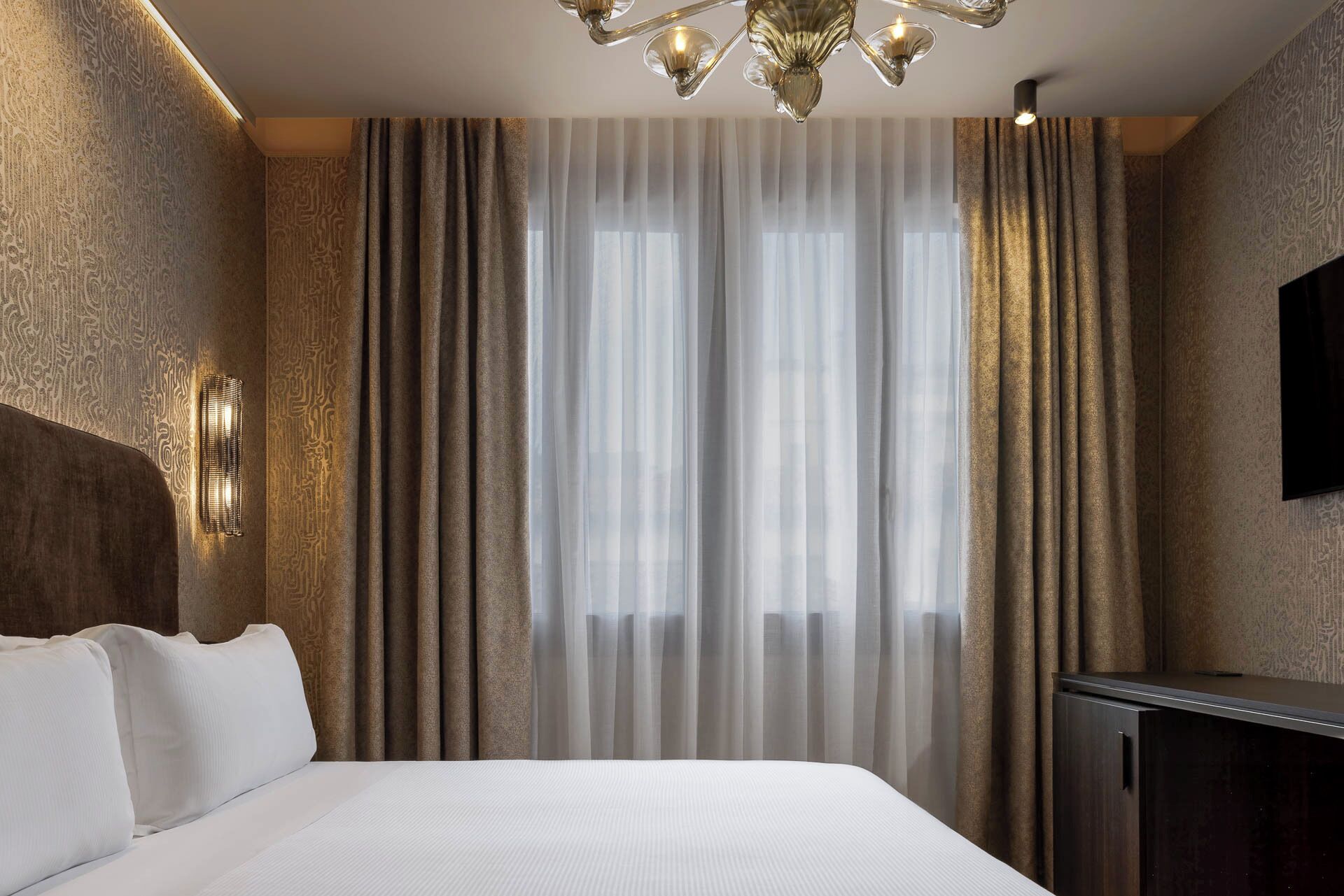 Quarto de hotel com uma cama grande, uma TV fixada na parede e uma janela com cortinas leves.