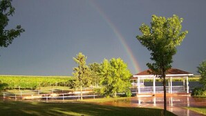 Ein Gazebo mit weißer Brüstung, umgeben von einer Rasenfläche und Bäumen, mit einem Weinberg im Hintergrund und einem Regenbogen am Himmel.