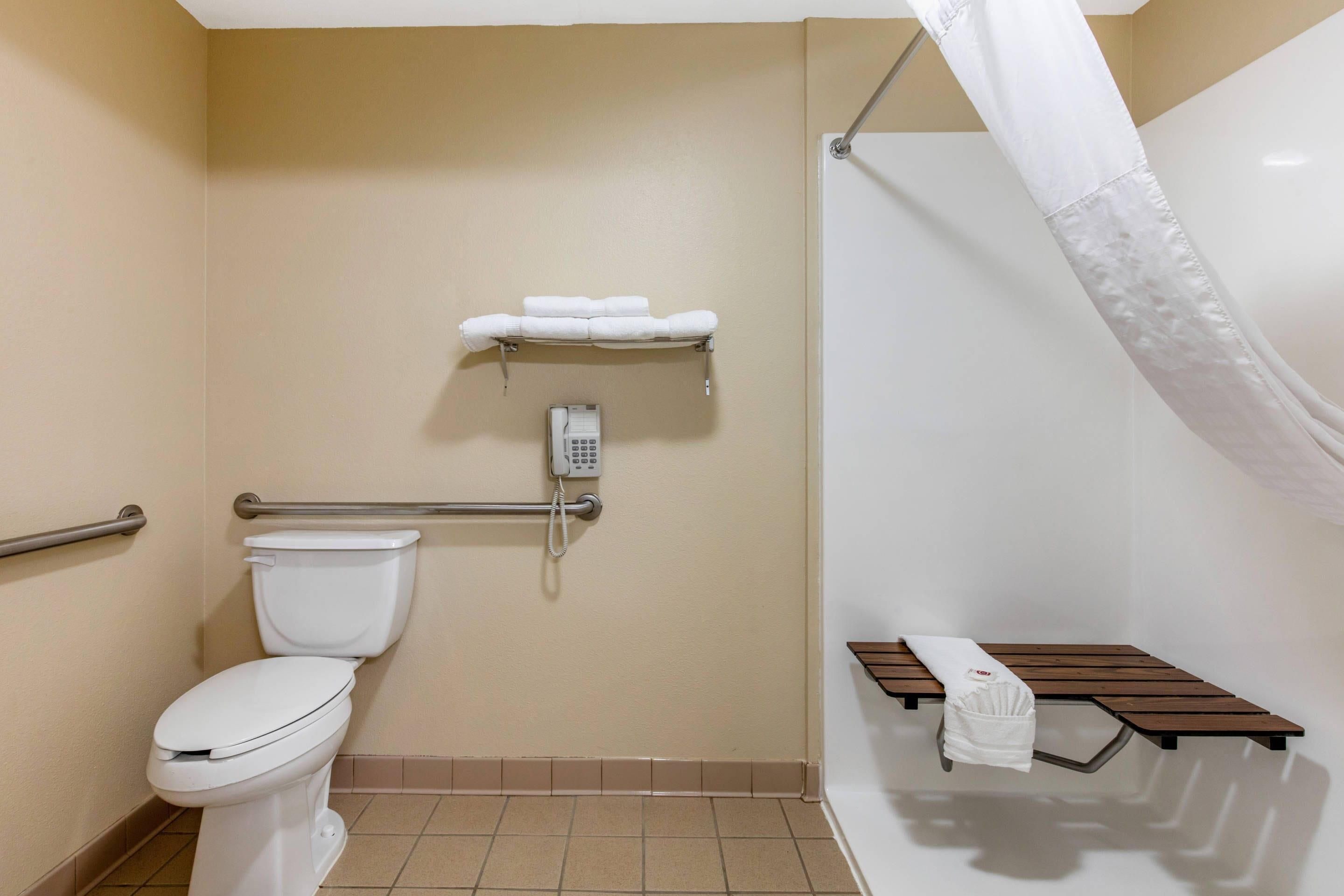 Une salle de bain équipée de toilettes, d’une douche avec un rideau blanc, d’une barre d’appui, d’une étagère avec des serviettes et d’un téléphone.