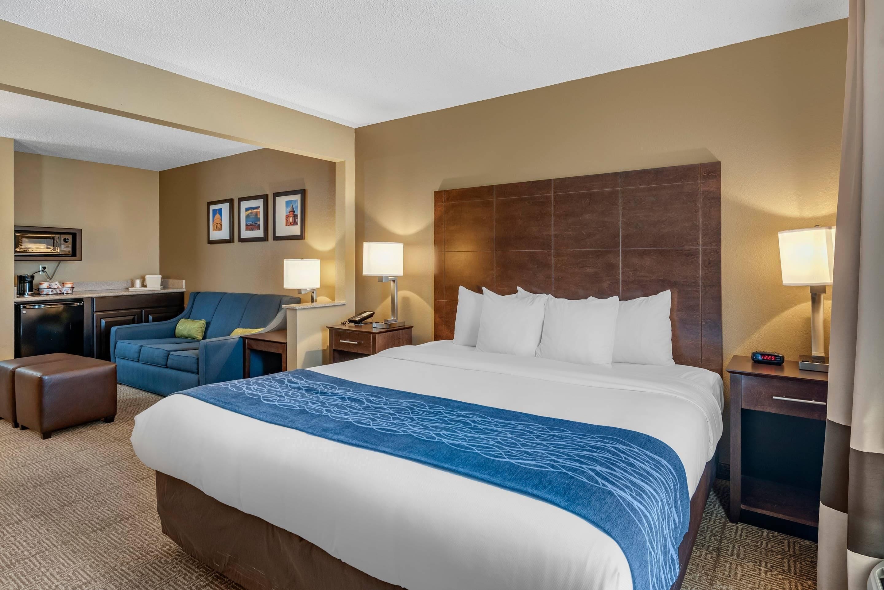 A hotel room with a large bed, a desk with a chair, a sofa, and a small kitchenette.