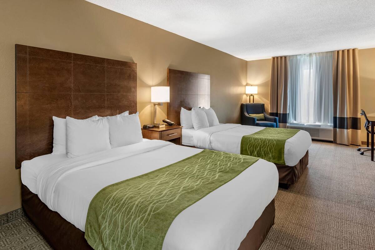 A hotel room with two beds, a desk, a chair, a lamp, and a large window with curtains.
