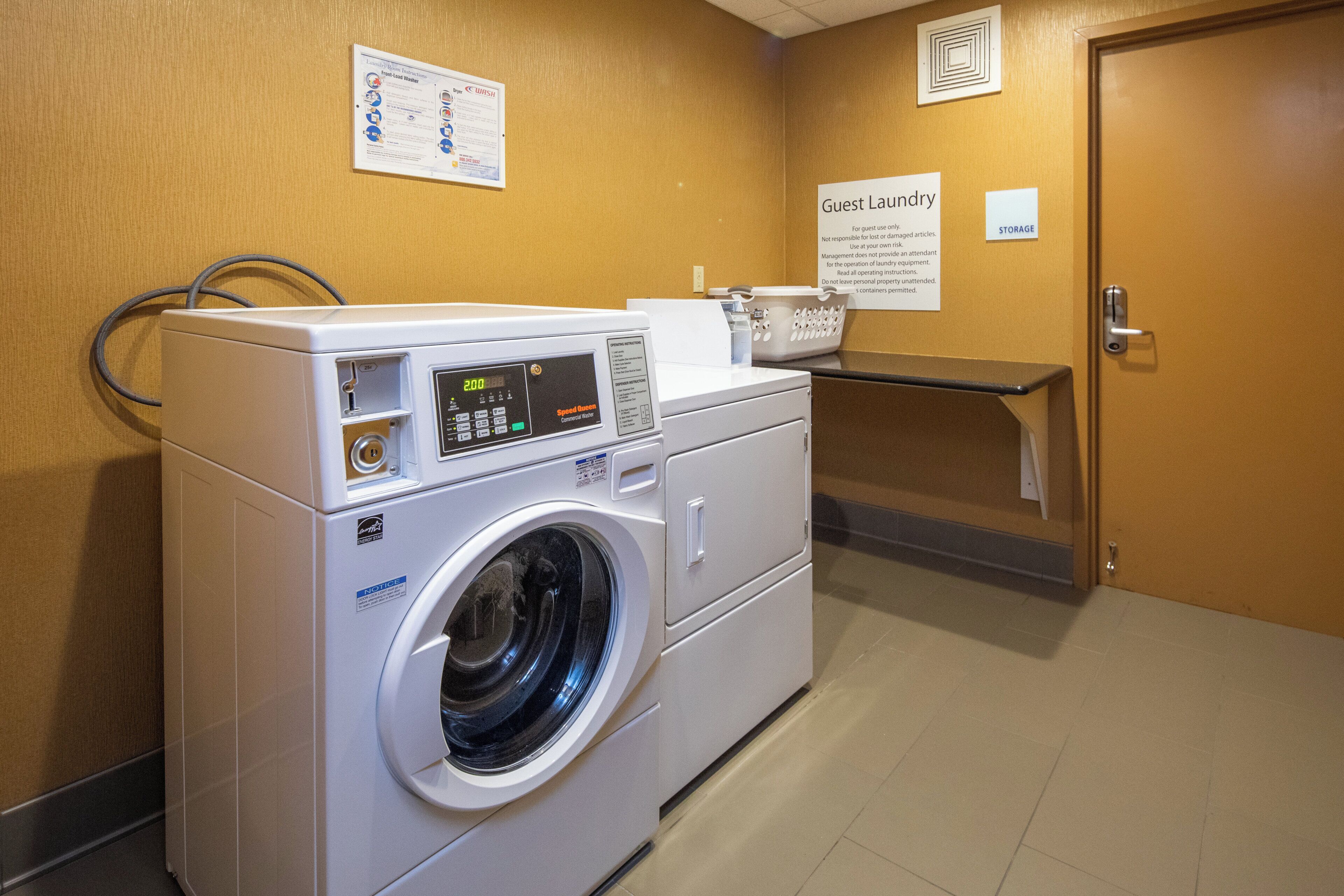 A front-load washing machine with a digital display, a wall-mounted laundry instruction sign, and a guest laundry storage area.