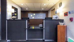A hotel reception desk with a digital clock, a fire alarm, and a sign indicating the availability of assistive devices. - Holiday Inn Express Castro Valley - I-580 by IHG (Castro Valley)