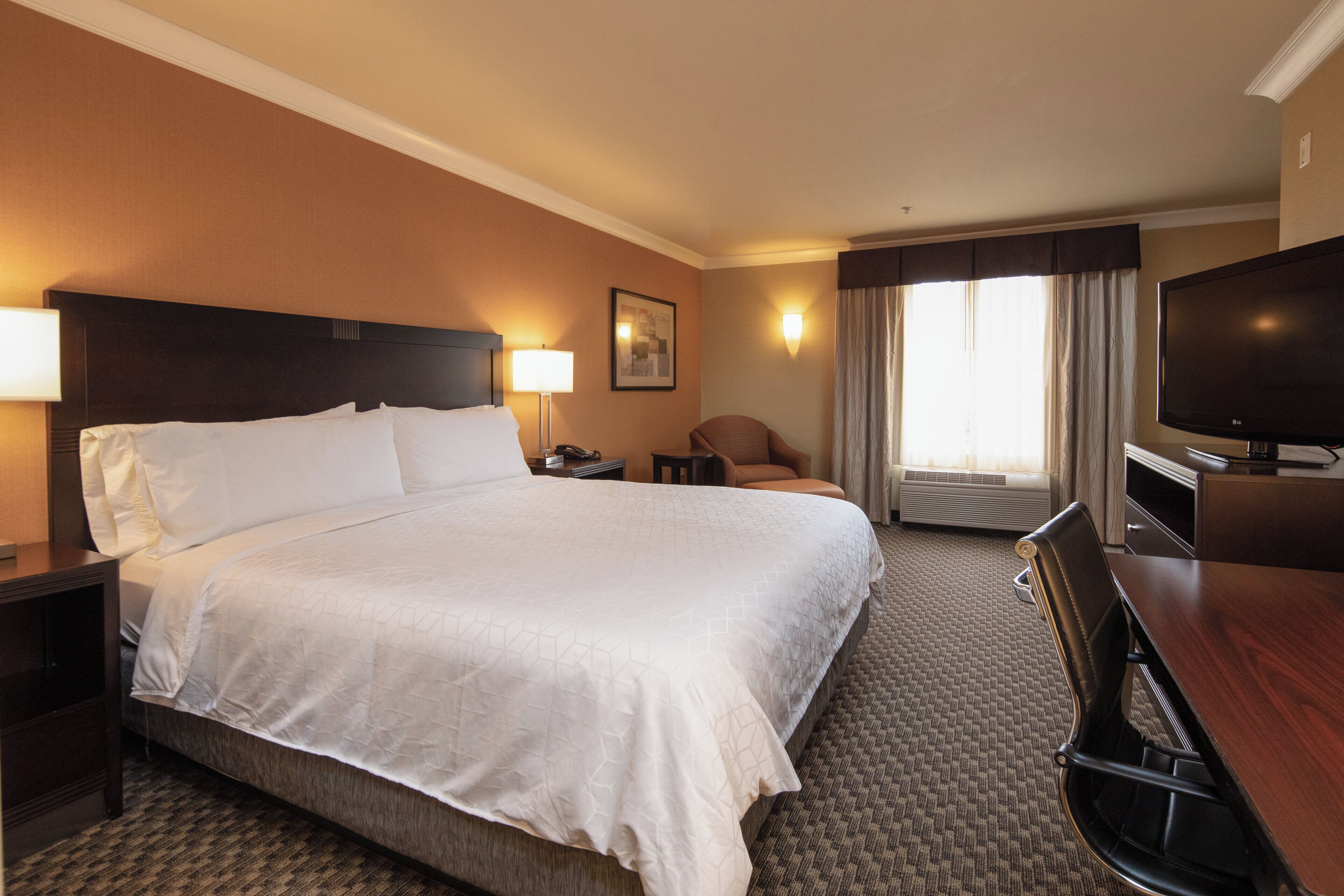 Une chambre d’hôtel avec un grand lit, un bureau, une chaise, une télévision et une fenêtre avec des rideaux.