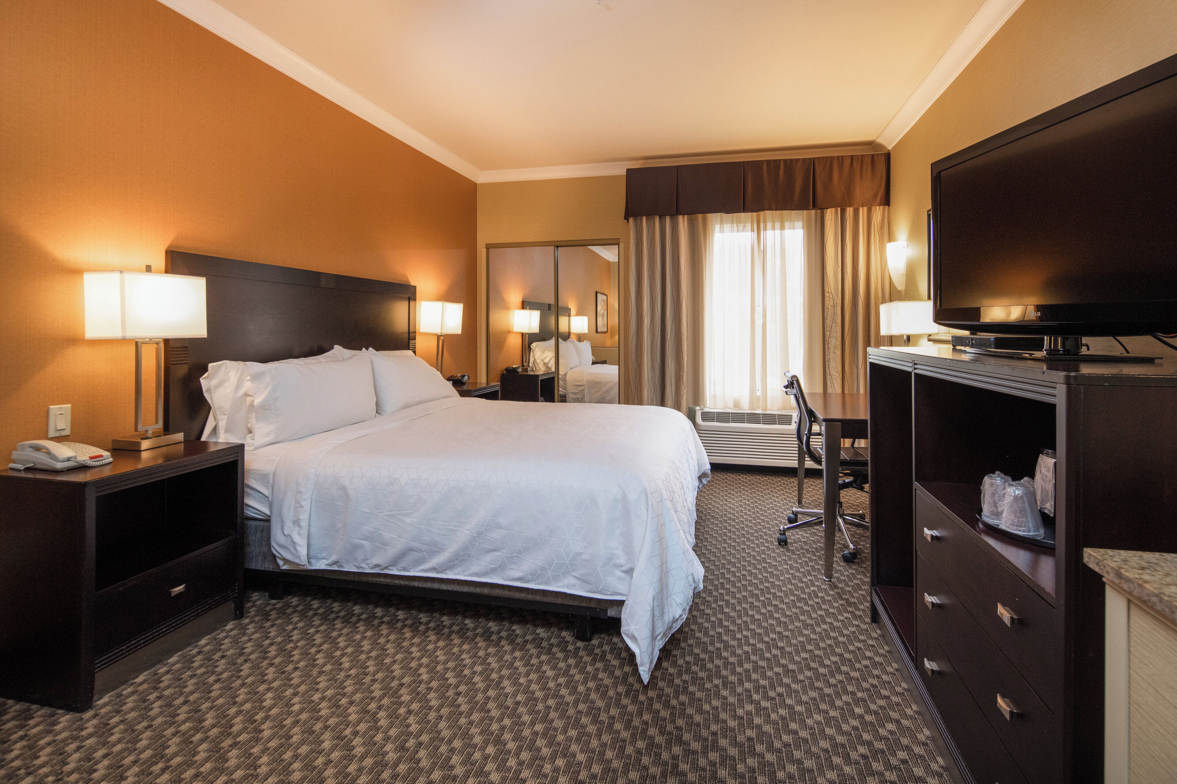 A hotel room with a large bed, bedside lamps, a desk with a computer, a television, and a mirror.