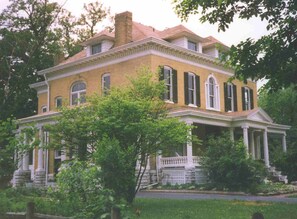 Une grande maison à deux étages, construite en briques, avec une façade symétrique, une entrée centrale flanquée de colonnes et un porche couvert.