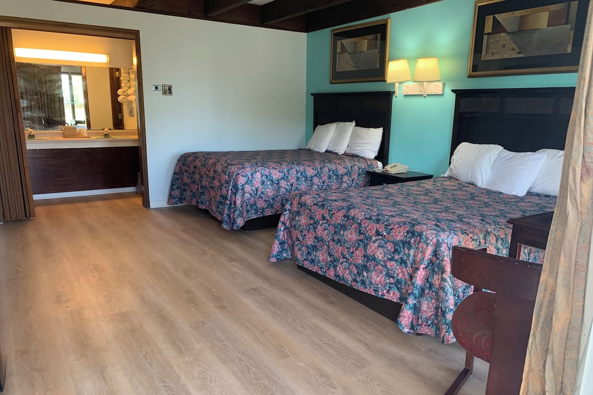 A room with two beds, wooden flooring, a bathroom with a mirror, and a door.
