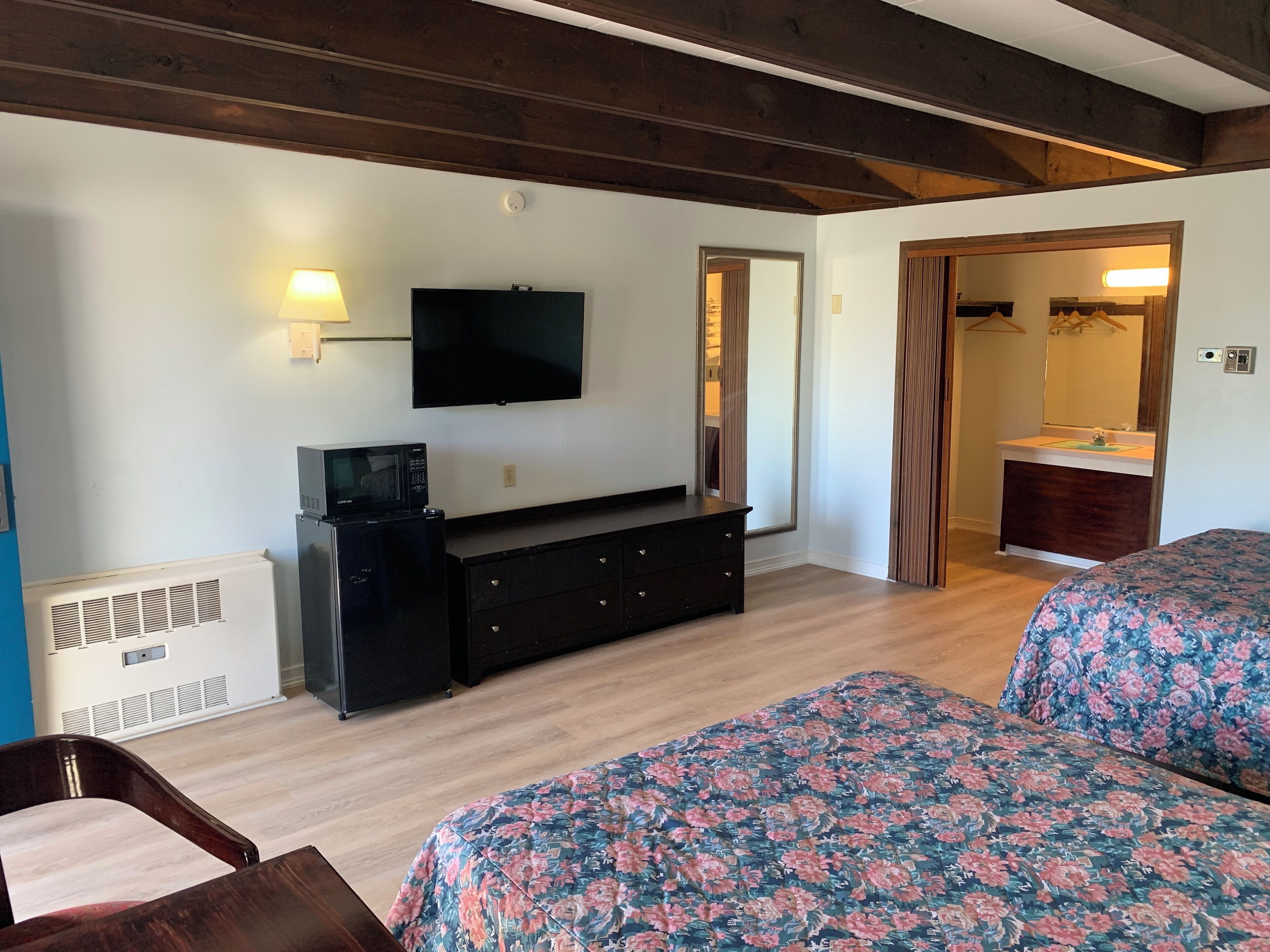 A hotel room with a flat-screen TV mounted on the wall, a dresser, a bed with floral bedding, a wooden floor, and a door leading to a bathroom.