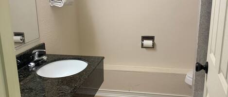 A bathroom with a granite countertop sink, a framed landscape painting on the wall, and a towel rack with folded towels.