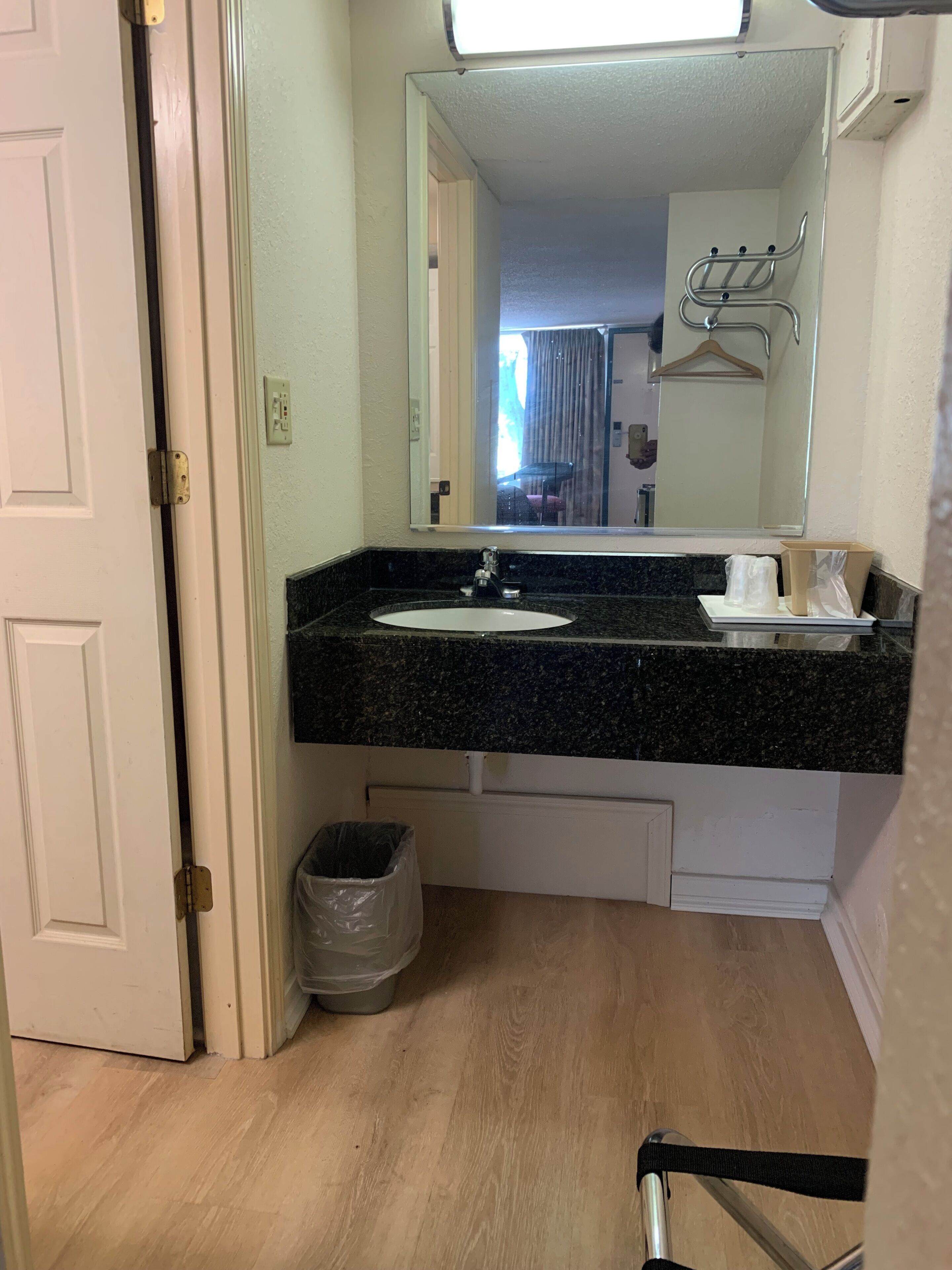 A bathroom with a granite countertop, a white sink, and a mirror above it. There is a trash can under the sink and a door leading to another room.