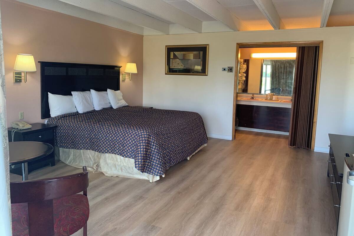 A hotel room with a large bed, bedside lamps, a desk with a telephone, a chair, and a bathroom visible through an open door.
