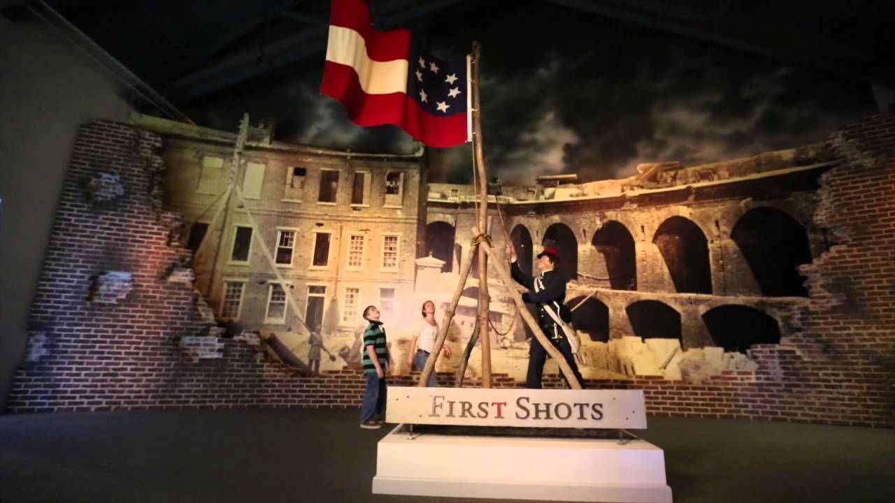A historical exhibit with a sign reading "First Shots," featuring a model of a building with a flag and a figure in uniform.