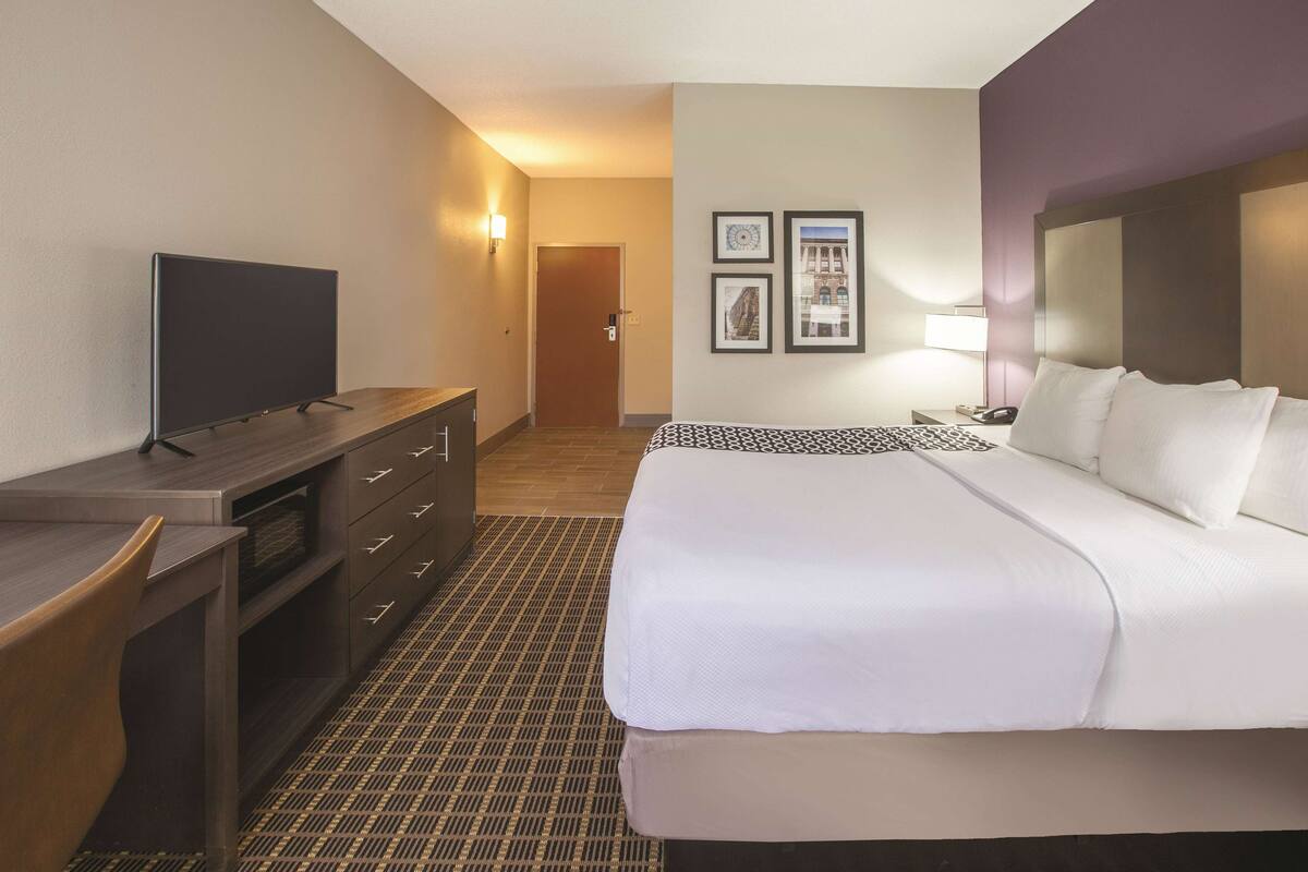 Une chambre d’hôtel avec un lit, un bureau doté d’un téléviseur à écran plat, une commode, un tapis à motifs et des tableaux encadrés accrochés au mur.