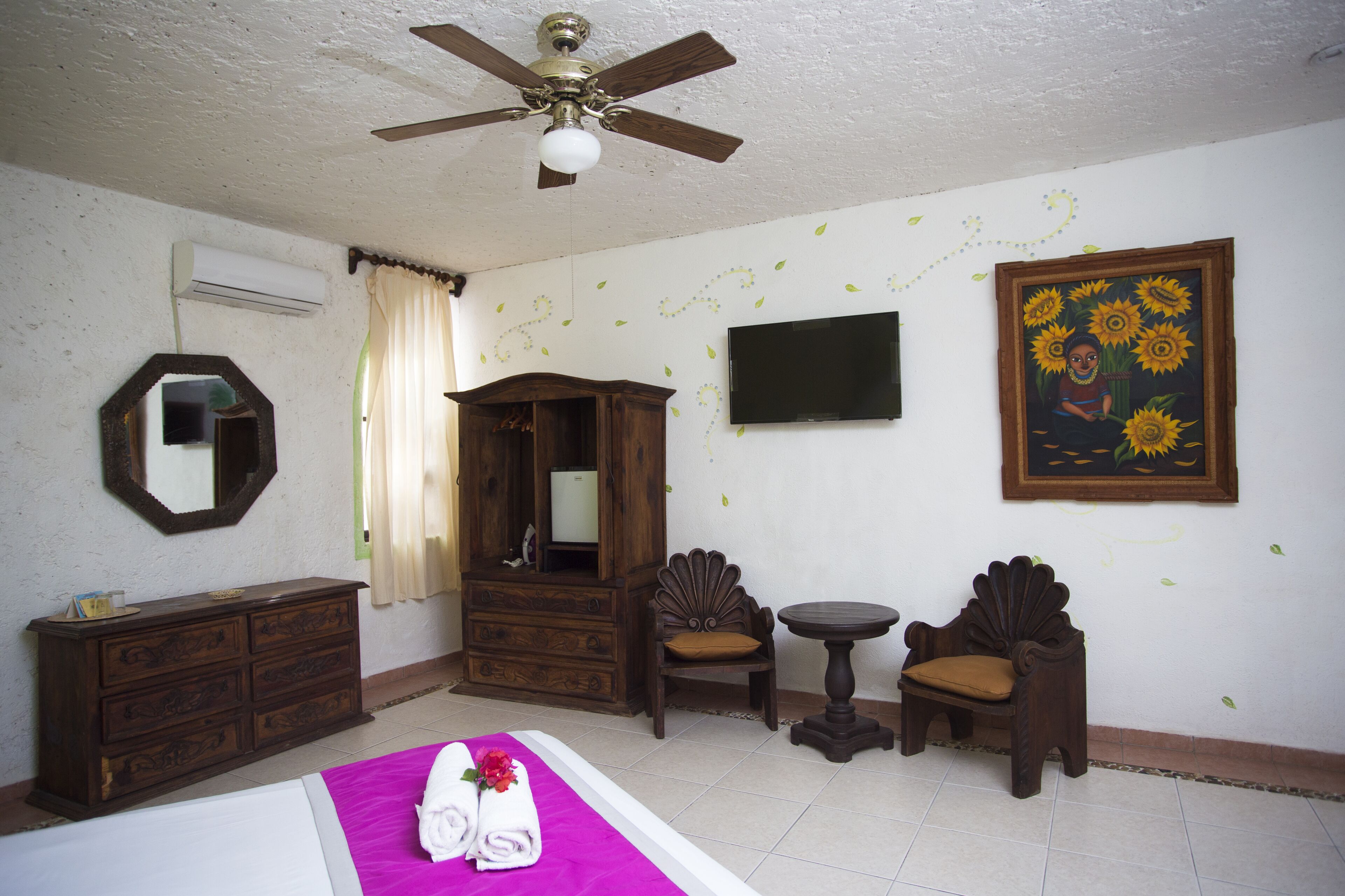 A hotel room with a bed, a wooden dresser, a vanity, a small table, two chairs, a ceiling fan, and a TV on the wall.