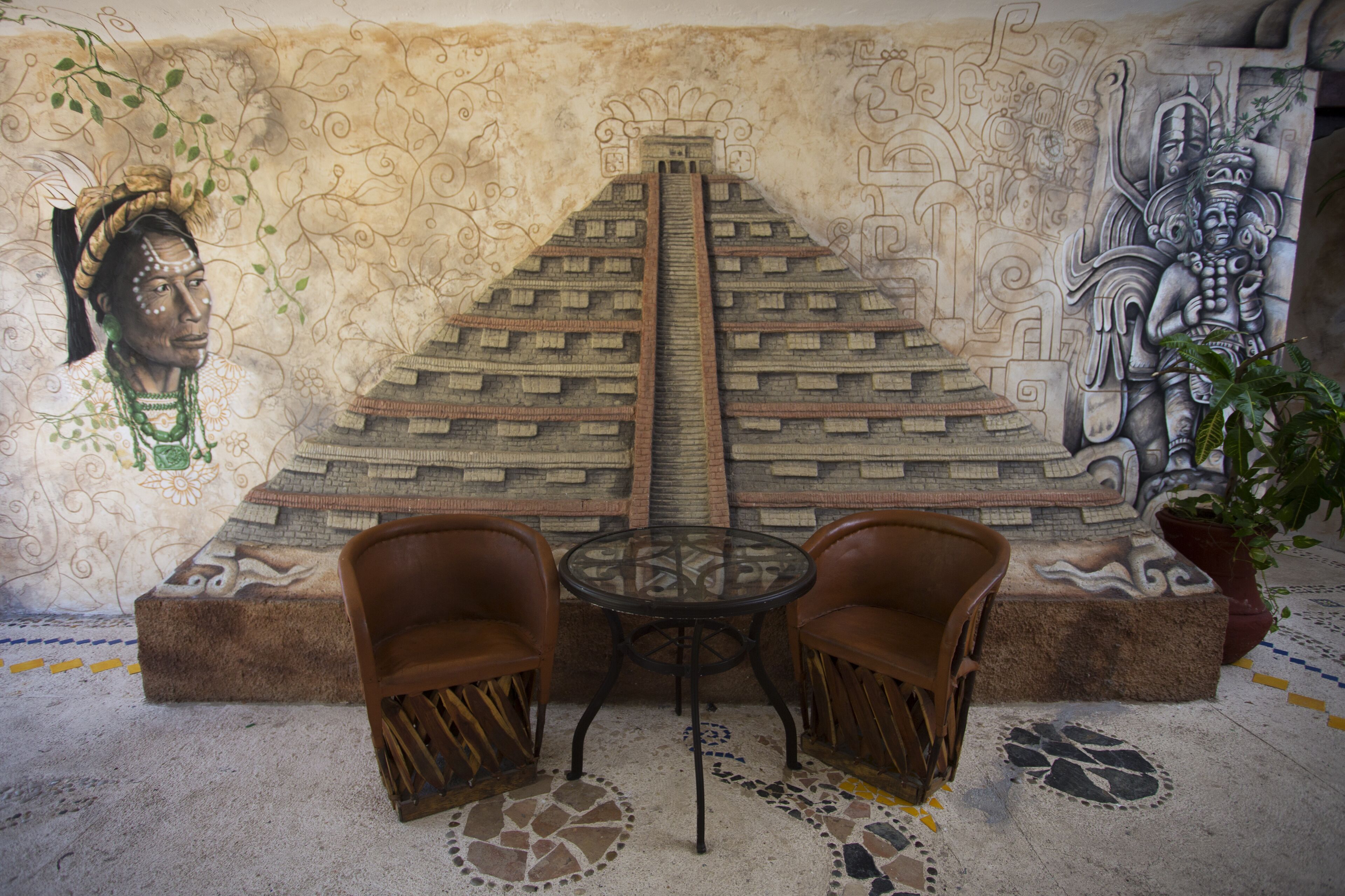 A mural of a pyramid and a seated figure, a round table, two chairs, and a potted plant.
