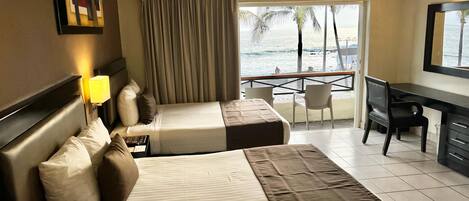 A hotel room with two beds, a desk, and a balcony with a view of the sea.