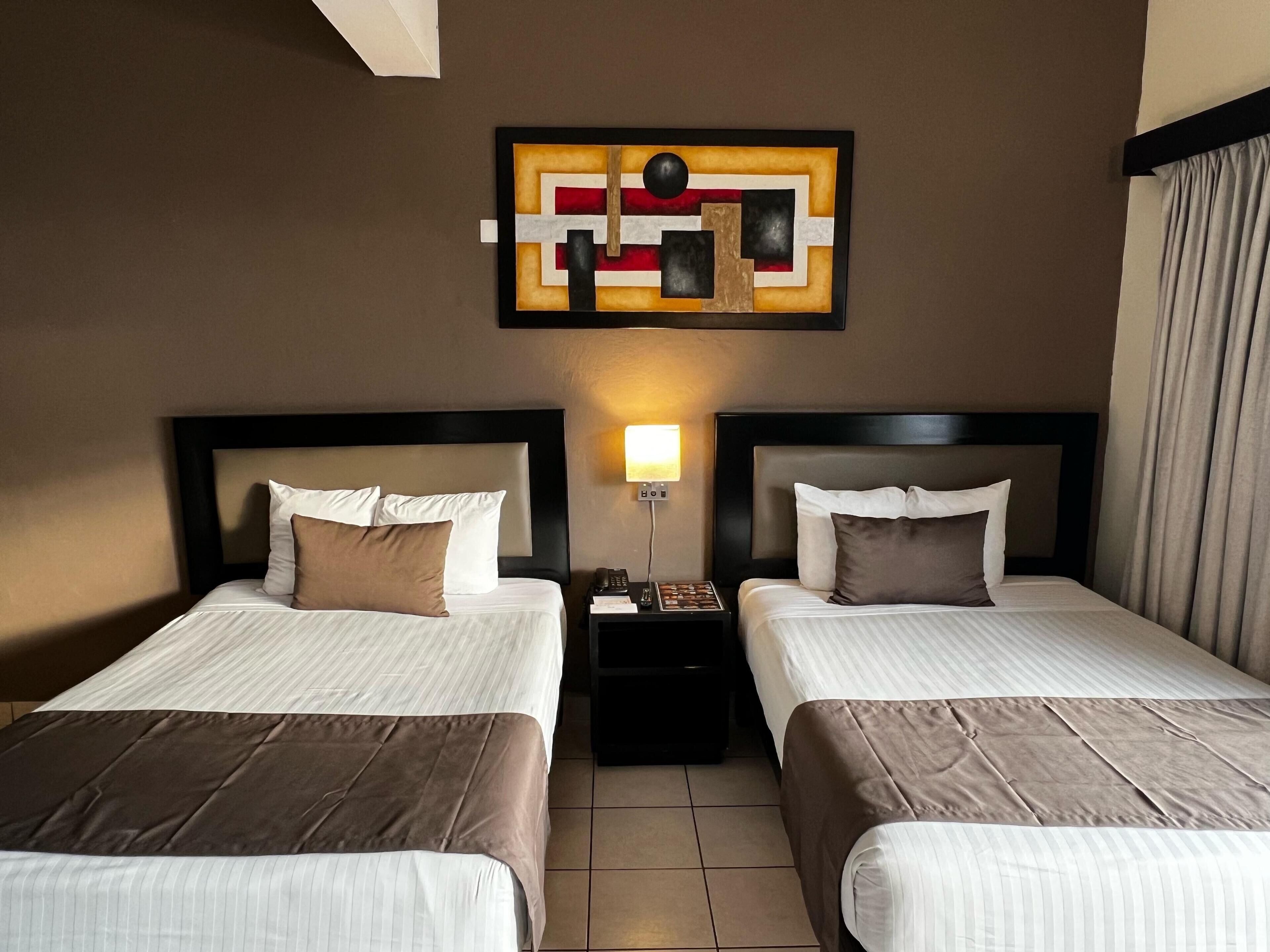 A hotel room with two beds, a nightstand with a lamp, and a wall-mounted abstract artwork.