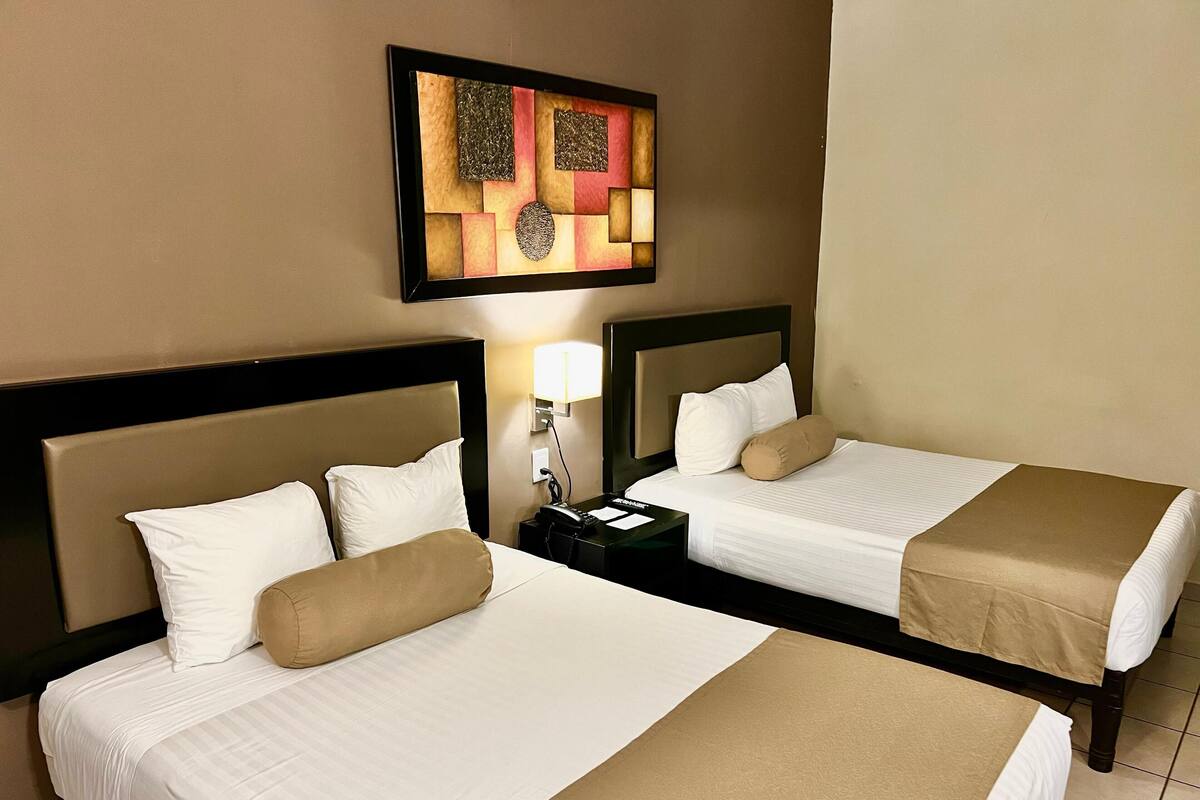A hotel room with two beds, each with white linens and beige throws, a nightstand with a lamp, and a wall-mounted artwork.