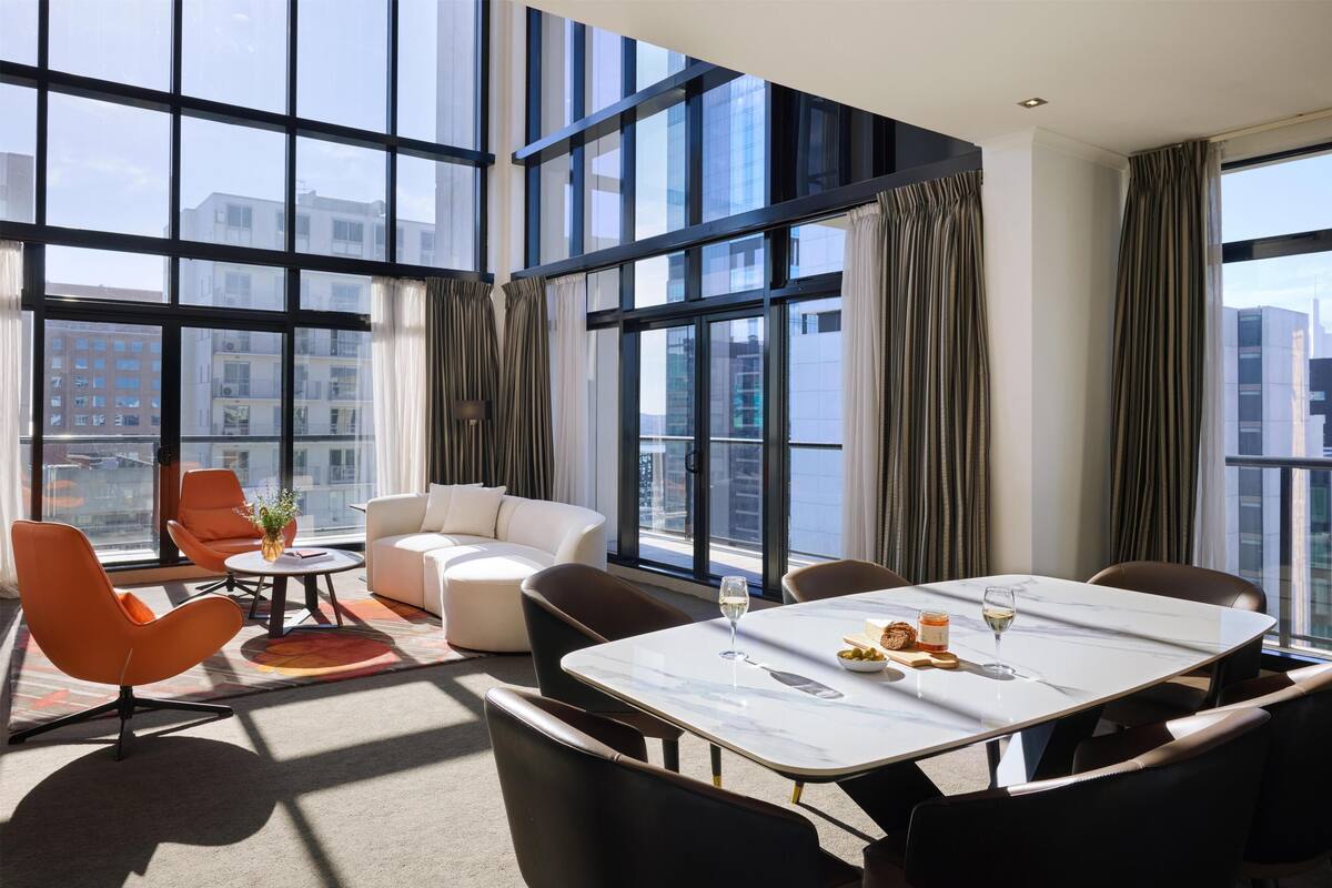 A modern hotel room with large windows, a dining area, and a seating arrangement.